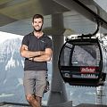 Ein lächelnder Mann in einem schwarzen T-Shirt steht mit verschränkten Armen vor einer Seilbahn. Im Hintergrund sind hohe Berge und ein klarer Himmel zu sehen.