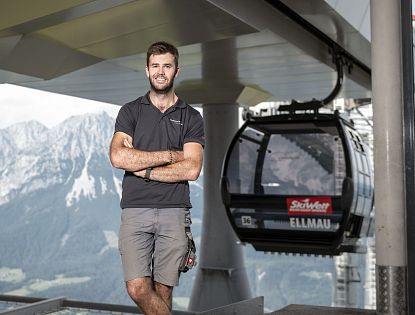 Ein lächelnder Mann in einem schwarzen T-Shirt steht mit verschränkten Armen vor einer Seilbahn. Im Hintergrund sind hohe Berge und ein klarer Himmel zu sehen.