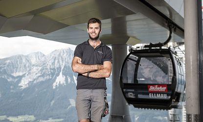 Ein lächelnder Mann in einem schwarzen T-Shirt steht mit verschränkten Armen vor einer Seilbahn. Im Hintergrund sind hohe Berge und ein klarer Himmel zu sehen.
