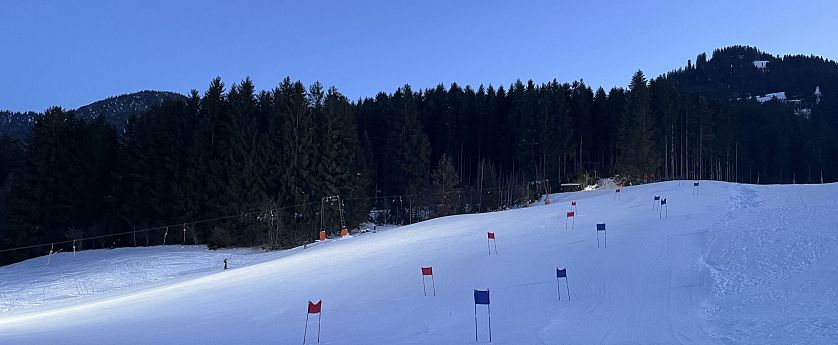 Sneeuwhelling omgeven door een dicht bos bij schemering, met blauwe en rode vlaggen langs het parcours, onder een heldere hemel.