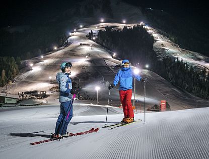 Zwei Skifahrer stehen auf präparierter Piste im beleuchteten Nachtskigebiet Söll. Im Hintergrund die beleuteten Skipisten zu sehen.