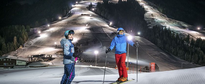 Zwei Skifahrer stehen auf präparierter Piste im beleuchteten Nachtskigebiet Söll. Im Hintergrund die beleuteten Skipisten zu sehen.