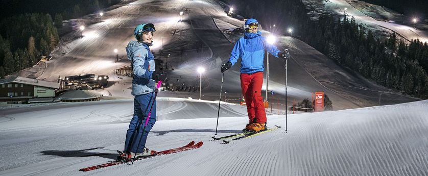 Zwei Skifahrer stehen auf präparierter Piste im beleuchteten Nachtskigebiet Söll. Im Hintergrund die beleuteten Skipisten zu sehen.