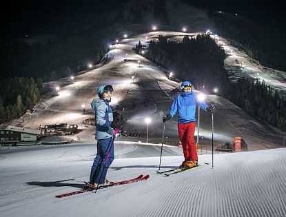 Zwei Skifahrer stehen auf präparierter Piste im beleuchteten Nachtskigebiet Söll. Im Hintergrund die beleuteten Skipisten zu sehen.