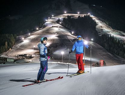 Zwei Skifahrer stehen auf präparierter Piste im beleuchteten Nachtskigebiet Söll. Im Hintergrund die beleuteten Skipisten zu sehen.