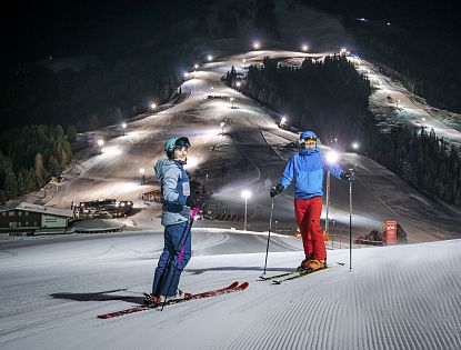 Zwei Skifahrer stehen auf präparierter Piste im beleuchteten Nachtskigebiet Söll. Im Hintergrund die beleuteten Skipisten zu sehen.