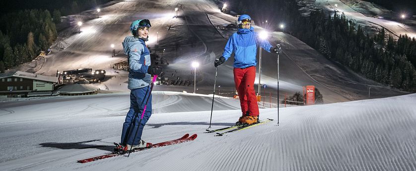 Zwei Skifahrer stehen auf präparierter Piste im beleuchteten Nachtskigebiet Söll. Im Hintergrund die beleuteten Skipisten zu sehen.