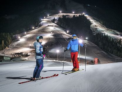 Zwei Skifahrer stehen auf präparierter Piste im beleuchteten Nachtskigebiet Söll. Im Hintergrund die beleuteten Skipisten zu sehen.