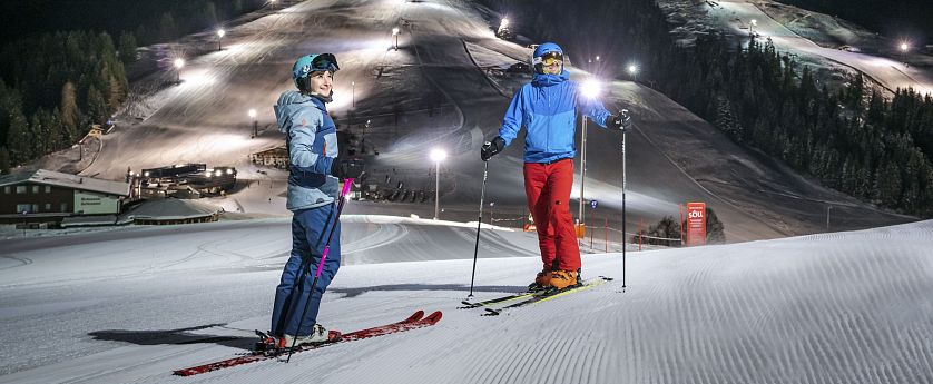 Zwei Skifahrer stehen auf präparierter Piste im beleuchteten Nachtskigebiet Söll. Im Hintergrund die beleuteten Skipisten zu sehen.