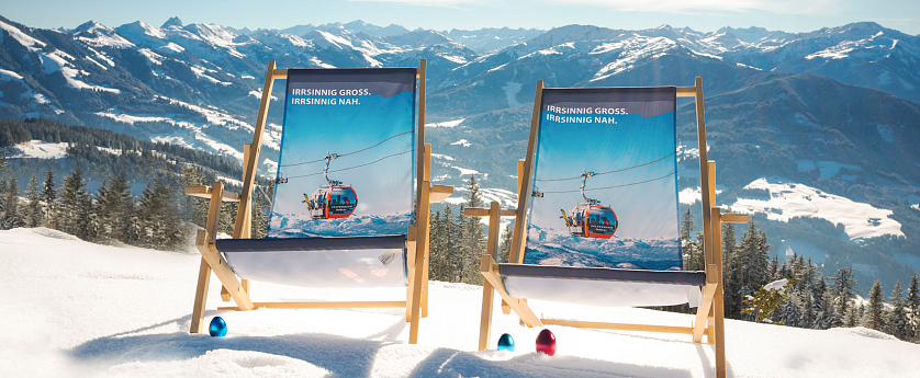 Zwei Liegestühle stehen im Schnee mit Blick auf eine Alpenlandschaft. Der Himmel ist klar und die Berge sind mit Schnee bedeckt.