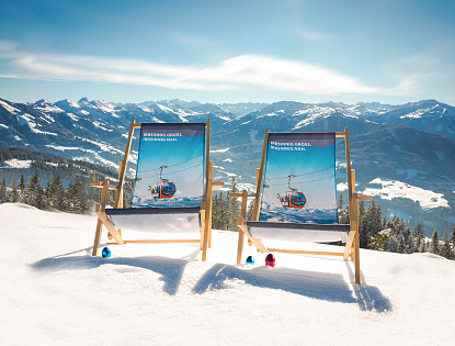 Zwei Liegestühle stehen im Schnee mit Blick auf eine Alpenlandschaft. Der Himmel ist klar und die Berge sind mit Schnee bedeckt.