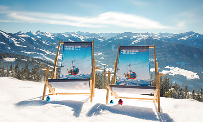 Zwei Liegestühle stehen im Schnee mit Blick auf eine Alpenlandschaft. Der Himmel ist klar und die Berge sind mit Schnee bedeckt.