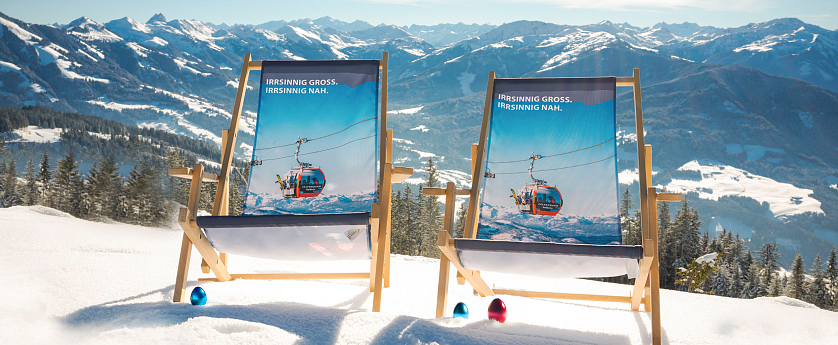Zwei Liegestühle stehen im Schnee mit Blick auf eine Alpenlandschaft. Der Himmel ist klar und die Berge sind mit Schnee bedeckt.