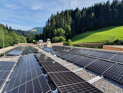 Solaranlagen auf einem Dach vor einer bewaldeten Berglandschaft unter blauem Himmel. Grüne Hügel und Bäume im Hintergrund.