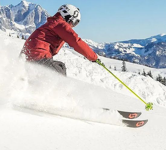 Ein Skifahrer in roter Jacke fährt einen schneebedeckten Hang hinunter, umgeben von Bergen und Bäumen unter einem klaren blauen Himmel.