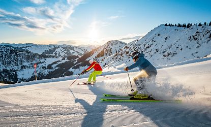 Zwei Skifahrer fahren in den Sonnenuntergang auf einer verschneiten Piste. Die Berge im Hintergrund sind schneebedeckt, und der Himmel strahlt in warmen Farben.