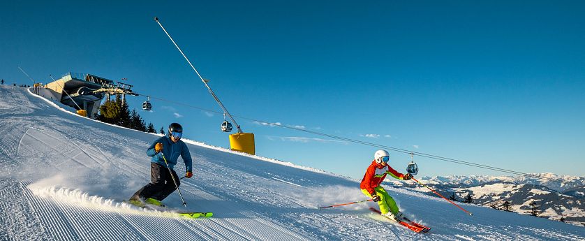 Zwei Skifahrer auf perfekt präparierter Piste in der SkiWelt bei Sonnenschein, dynamische Abfahrt mit Gondelbahn im Hintergrund.