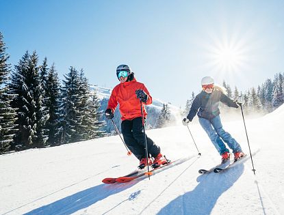 Zwei Skifahrer fahren auf einer verschneiten Piste. Die Sonne strahlt am blauen Himmel, umgeben von Tannenbäumen.
