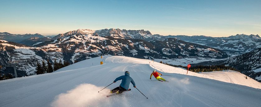Zwei Skifahrer gleiten die schneebedeckte Piste hinunter, umgeben von majestätischen Bergen. Der Himmel ist klar und blau, die Landschaft strahlt winterliche Ruhe aus.