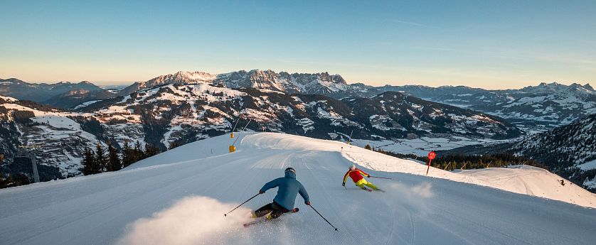 Zwei Skifahrer gleiten die schneebedeckte Piste hinunter, umgeben von majestätischen Bergen. Der Himmel ist klar und blau, die Landschaft strahlt winterliche Ruhe aus.