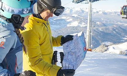 Ein Mann in gelber Ski-Jacke und eine Frau in blauer Jacke betrachten auf einer schneebedeckten Piste eine Karte. Im Hintergrund sind schneebedeckte Berge und ein Skilift zu sehen.