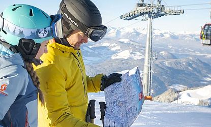 Zwei Personen in Skibekleidung studieren eine Karte auf einer verschneiten Berglandschaft mit Blick auf Skilifte und entfernte Berge.
