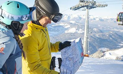 Zwei Personen in Skiausrüstung betrachten eine Karte in einer verschneiten Berglandschaft. Ein Skilift ist im Hintergrund zu sehen.