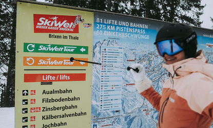 Eine Wintersportlerin steht vor einem großen Informationsschild mit einer Übersichtskarte der Skigebiete. Sie zeigt mit einem Stock auf die verschiedenen Pisten und Lifte.