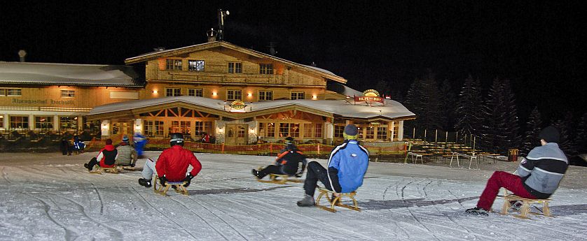 Menschen rodeln nachts auf einer beleuchteten Piste vor einem alpinen Gebäude im Schnee.