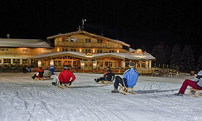 Menschen rodeln nachts auf einer beleuchteten Piste vor einem alpinen Gebäude im Schnee.