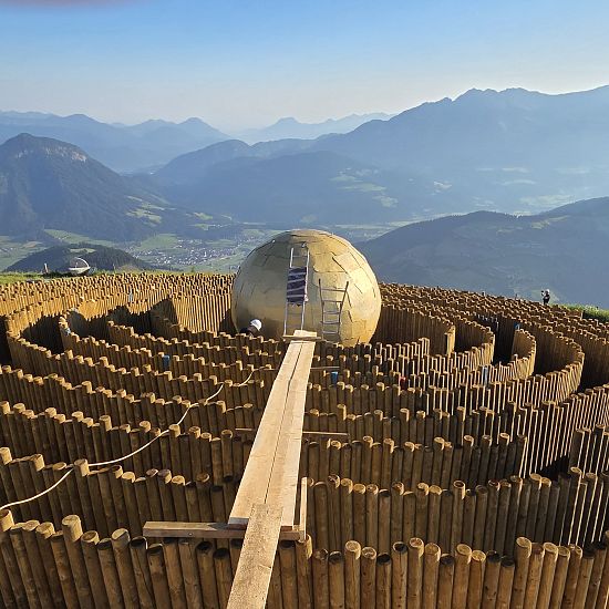 Ein Aussichtspunkt auf einem Berg präsentiert ein großes, kugelförmiges Objekt inmitten eines hölzernen Heckenlabyrinths, umgeben von Berglandschaft.