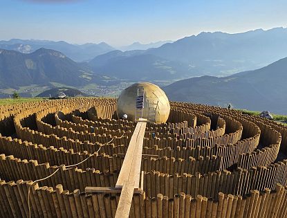Ein Aussichtspunkt auf einem Berg präsentiert ein großes, kugelförmiges Objekt inmitten eines hölzernen Heckenlabyrinths, umgeben von Berglandschaft.