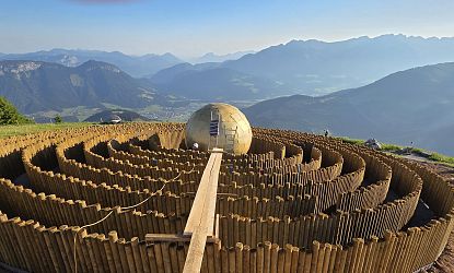 Ein Aussichtspunkt auf einem Berg präsentiert ein großes, kugelförmiges Objekt inmitten eines hölzernen Heckenlabyrinths, umgeben von Berglandschaft.