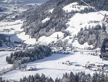 Verschneite Berglandschaft mit einer Seilbahn, die über eine winterliche Szenerie mit Wäldern, Hügeln und verstreuten Gebäuden fährt. Blauer Himmel im Hintergrund.