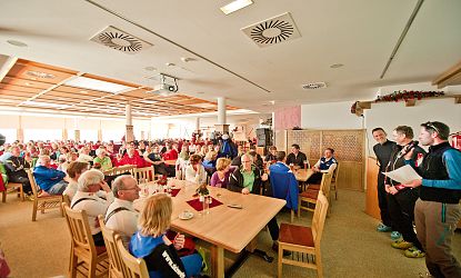 Eine große Gruppe von Menschen sitzt in einem holzvertäfelten Saal, während ein paar Personen vorne stehen und sprechen. Es herrscht eine entspannte Atmosphäre.