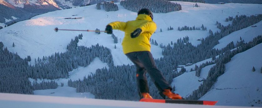 Skifahrer mit gelber Jacke fährt Talabwärts bei Sonnenaufgang. Im Hintergrund mit ersten Sonnenstrahlen beleuteter Wilde Kaiser.