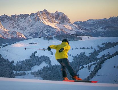 Skifahrer mit gelber Jacke fährt Talabwärts bei Sonnenaufgang. Im Hintergrund mit ersten Sonnenstrahlen beleuteter Wilde Kaiser.