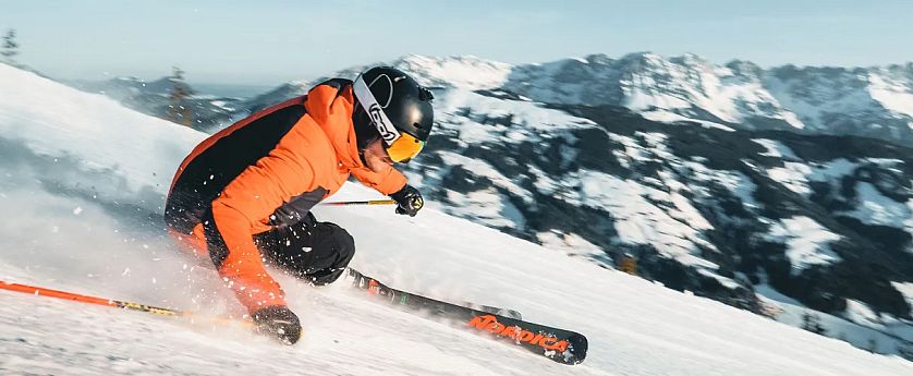 Muž ve sportovní výbavě sjíždí na lyžích zasněžený svah s horskou krajinou v pozadí. Bílý sníh, modrá obloha a muž v oranžové bundě s helmou.