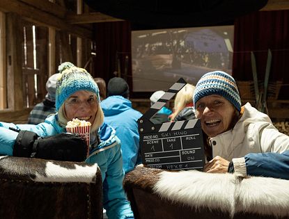 Zwei Personen sitzen in einer rustikalen Umgebung, beide lächeln in die Kamera. Eine Person hält eine Tüte Popcorn, während die andere eine Klappe mit Filmdetails präsentiert. Im Hintergrund ist ein Projektor sichtbar.