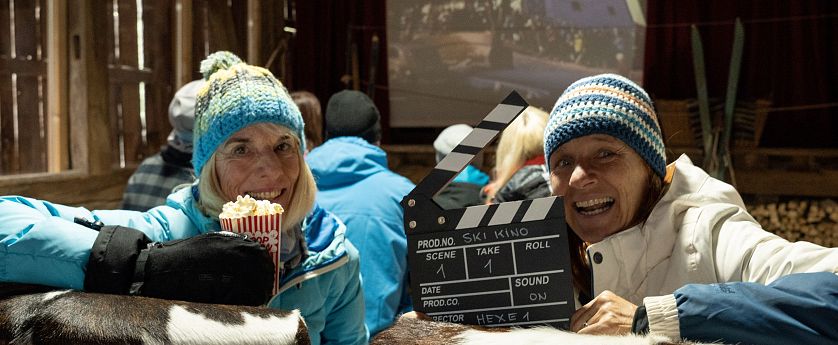 Zwei Personen sitzen in einer rustikalen Umgebung, beide lächeln in die Kamera. Eine Person hält eine Tüte Popcorn, während die andere eine Klappe mit Filmdetails präsentiert. Im Hintergrund ist ein Projektor sichtbar.