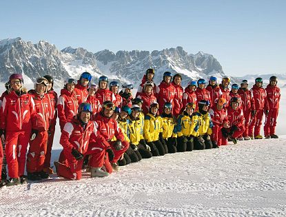 Eine Gruppe von Skiinstruktoren posiert im Schnee vor einer Berglandschaft. Die Instruktoren tragen rote und gelbe Skikleidung, die Berge im Hintergrund sind schnee-covered.