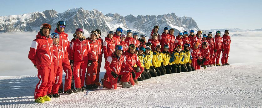 Eine Gruppe von Skiinstruktoren posiert im Schnee vor einer Berglandschaft. Die Instruktoren tragen rote und gelbe Skikleidung, die Berge im Hintergrund sind schnee-covered.