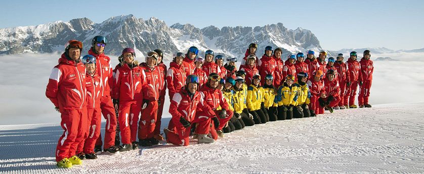 Eine Gruppe von Skiinstruktoren posiert im Schnee vor einer Berglandschaft. Die Instruktoren tragen rote und gelbe Skikleidung, die Berge im Hintergrund sind schnee-covered.