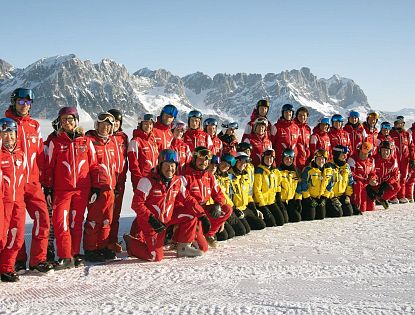 Eine Gruppe von Skiinstruktoren posiert im Schnee vor einer Berglandschaft. Die Instruktoren tragen rote und gelbe Skikleidung, die Berge im Hintergrund sind schnee-covered.