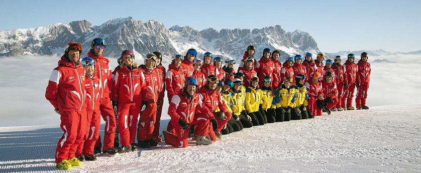 Eine Gruppe von Skiinstruktoren posiert im Schnee vor einer Berglandschaft. Die Instruktoren tragen rote und gelbe Skikleidung, die Berge im Hintergrund sind schnee-covered.
