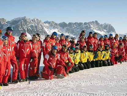 Eine Gruppe von Skiinstruktoren posiert im Schnee vor einer Berglandschaft. Die Instruktoren tragen rote und gelbe Skikleidung, die Berge im Hintergrund sind schnee-covered.