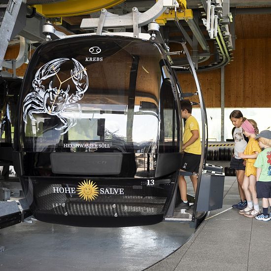 Group of people waiting to board a cable car at a station, with a black gondola marked with a scorpion logo.