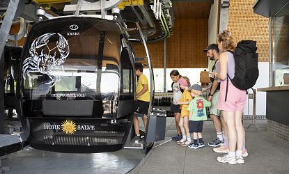 Group of people waiting to board a cable car at a station, with a black gondola marked with a scorpion logo.