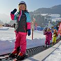 Ein kleines Kind steht auf Skiern und hält den Skistock hoch, während es auf einem Förderband im Schnee steht. Im Hintergrund sind weitere Kinder und verschneite Berge zu sehen.