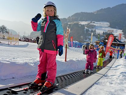Ein kleines Kind steht auf Skiern und hält den Skistock hoch, während es auf einem Förderband im Schnee steht. Im Hintergrund sind weitere Kinder und verschneite Berge zu sehen.